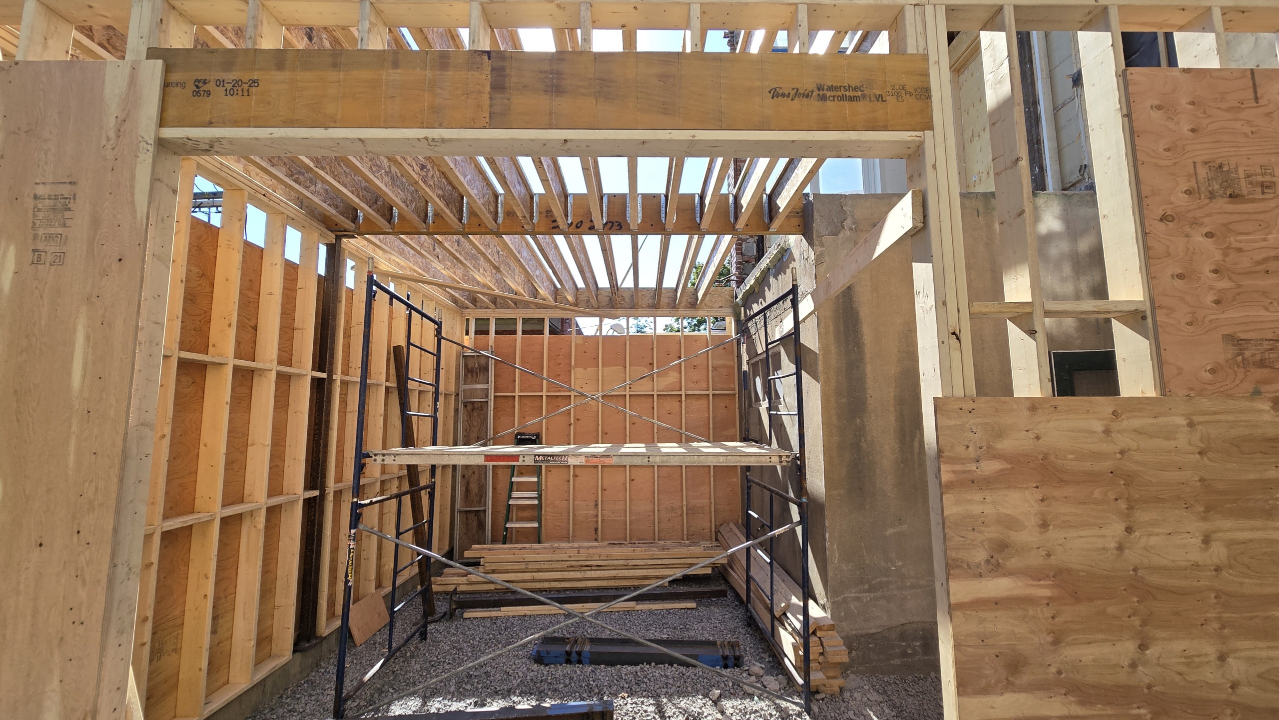 Interior of a wood-framed building under construction, with vertical studs, ceiling joists, and metal scaffolding in the center
