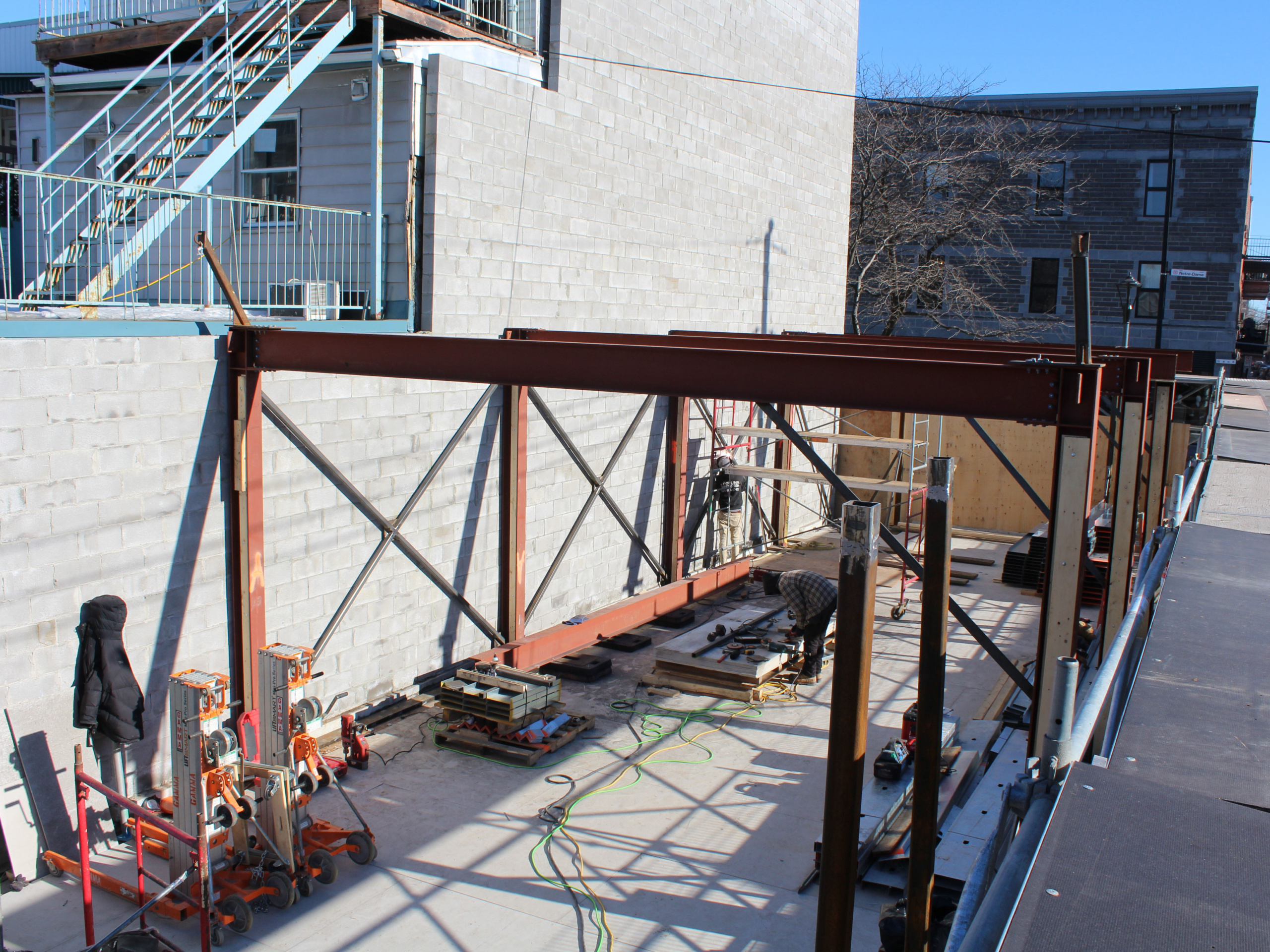 Construction area with orange steel framework and scaffolding beside a block-building under construction; tools and hoses on the ground with workers nearby.