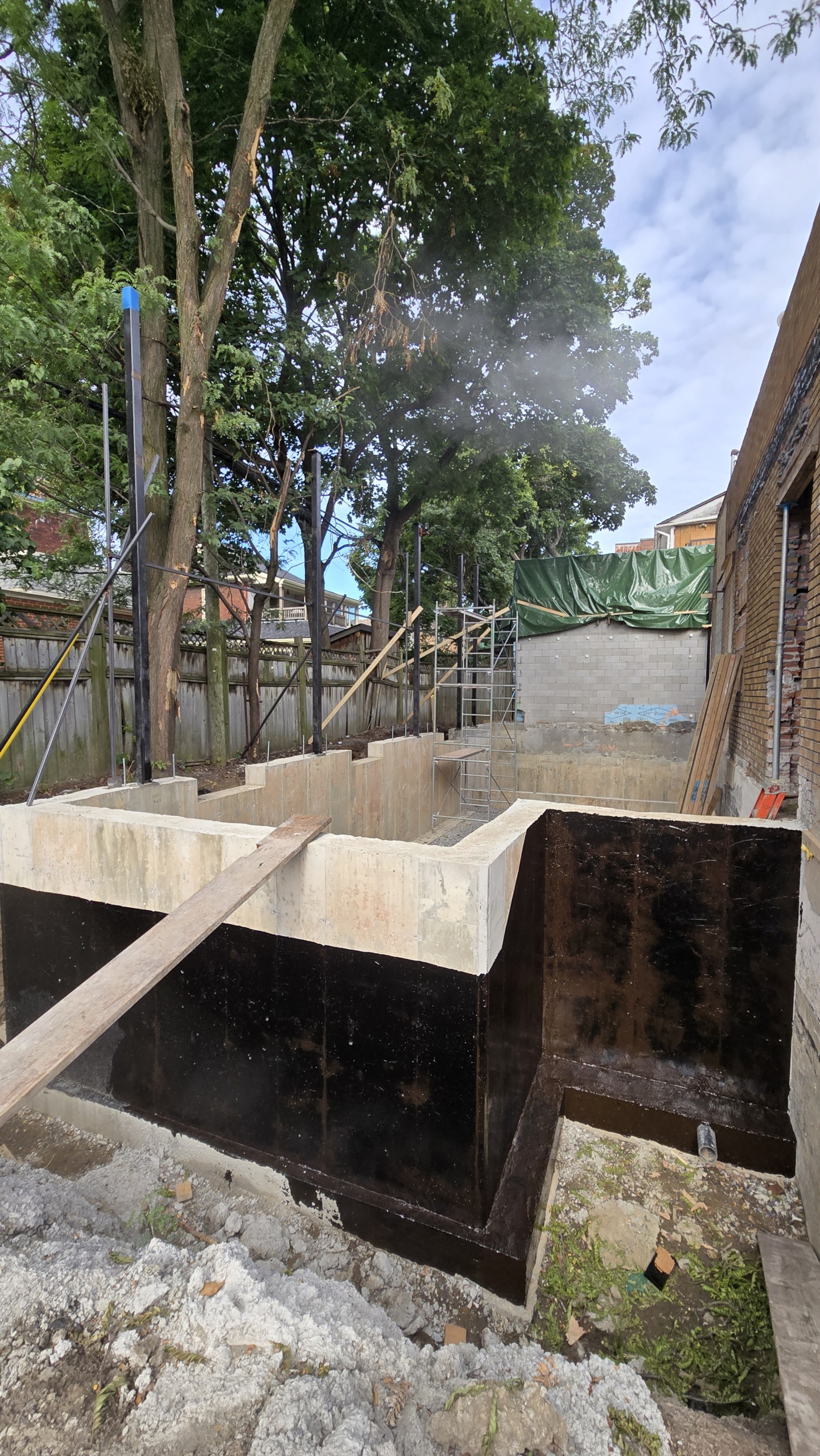 Deep rectangular foundation pit with black waterproof concrete walls, scaffolding, and wooden planks at a construction site