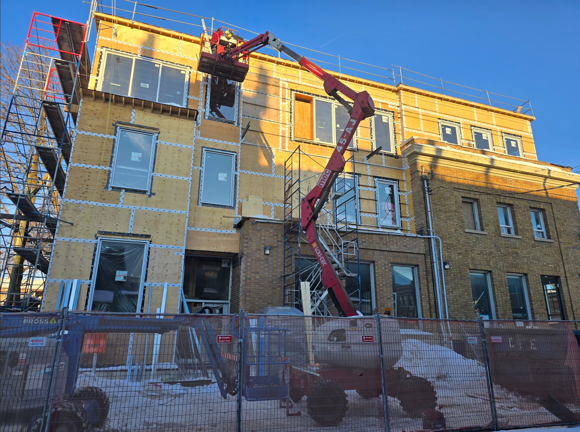 Construction site: multi-story building under renovation with plywood sheathing, scaffolding, and a red boom lift extending to the upper floor against a blue sky.