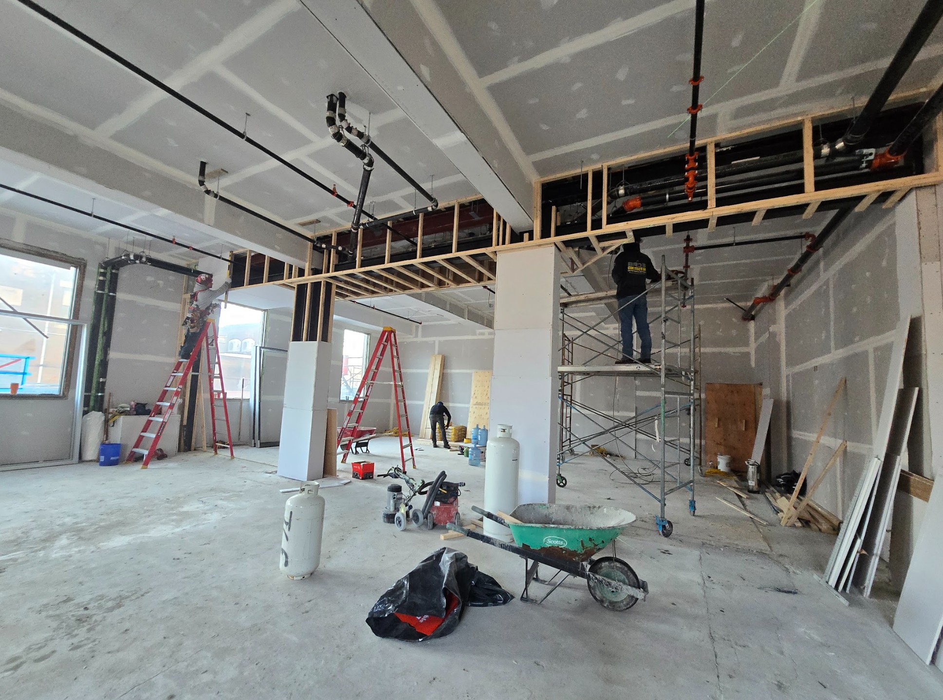 Construction site interior with exposed framing, scaffolding, and workers installing piping and drywall. Red ladders and construction tools scattered on the concrete floor.