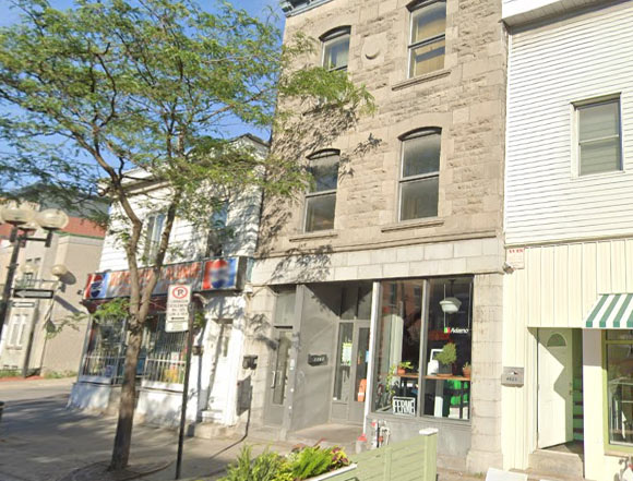 Urban storefront on a three-story stone building with glass entry and large display windows, shaded by a tree on the sidewalk.