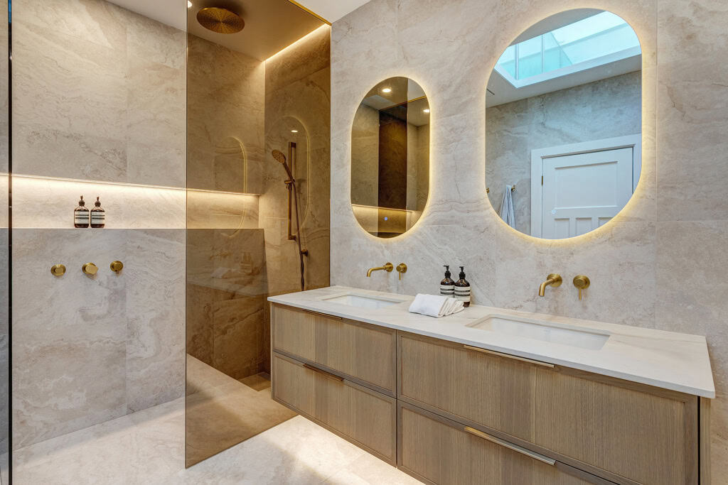 Luxurious marble bathroom with a double-sink vanity, round backlit mirrors, and brass fixtures across a light wood cabinet.