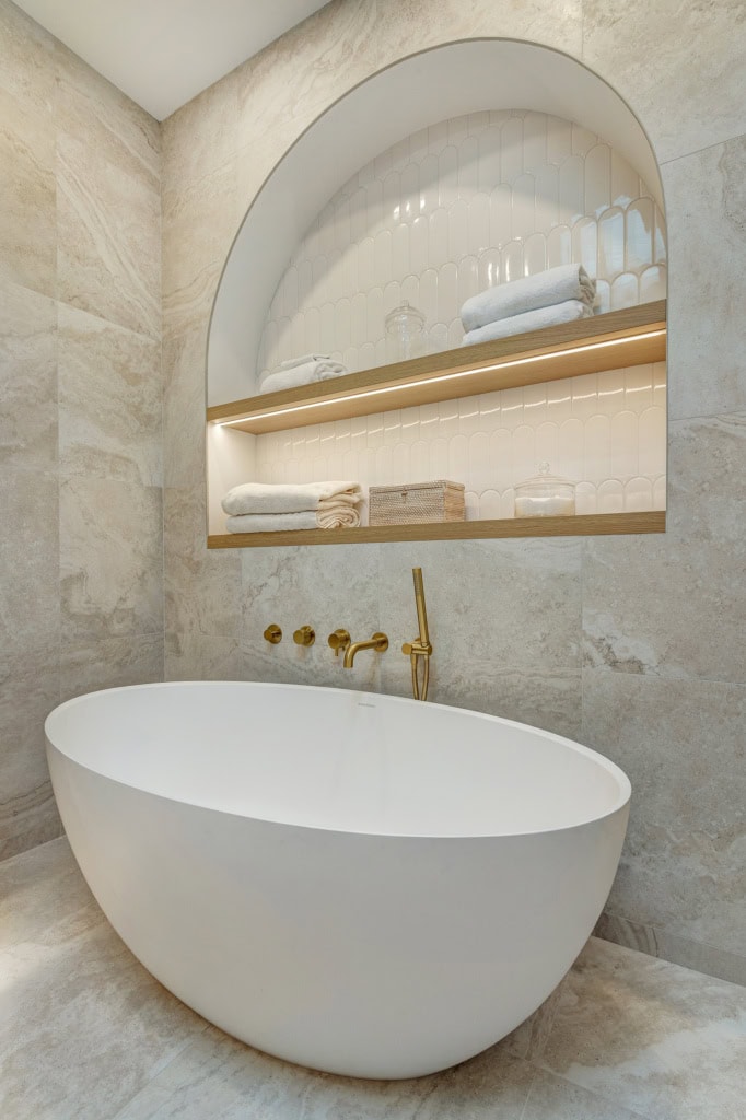 White freestanding bathtub with brushed gold fixtures in a marble-tiled bathroom, arched shelf niche above with towels and glass jar