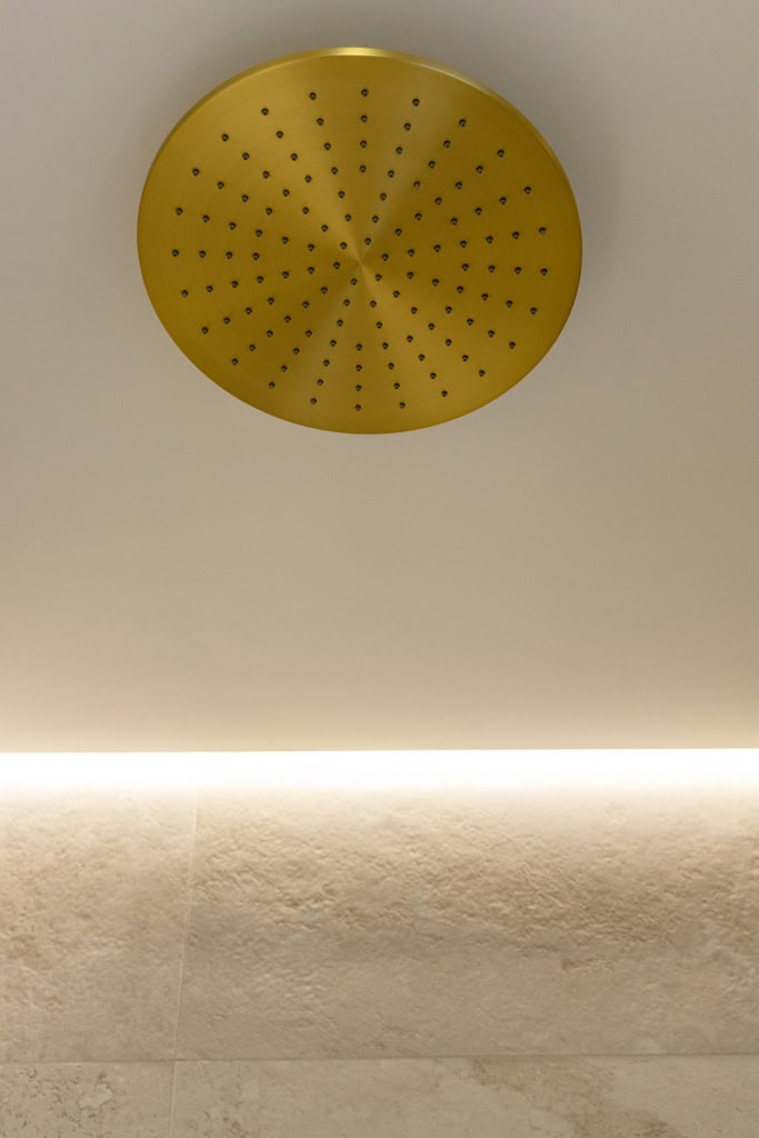 Gold circular ceiling shower head mounted on a beige bathroom ceiling in overhead view.