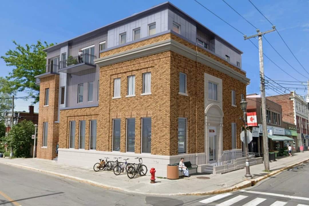Construction site: multi-story building under renovation with plywood sheathing, scaffolding, and a red boom lift extending to the upper floor against a blue sky.