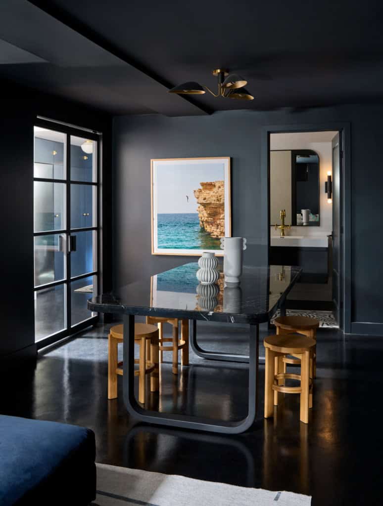 Dark modern dining area with a black marble table, light wood stools, and a seaside framed print on the wall.